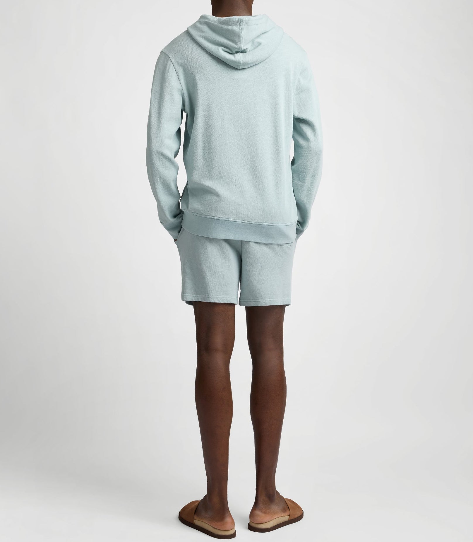 Person stands with back to camera wearing a light blue hoodie, Onia Slub Terry Short, and brown slide sandals against a plain background.