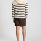 A person stands facing away against a plain background, wearing the Onia Frank Hooded Sweater—a cream and black striped breathable knit—paired with brown shorts and brown sandals.