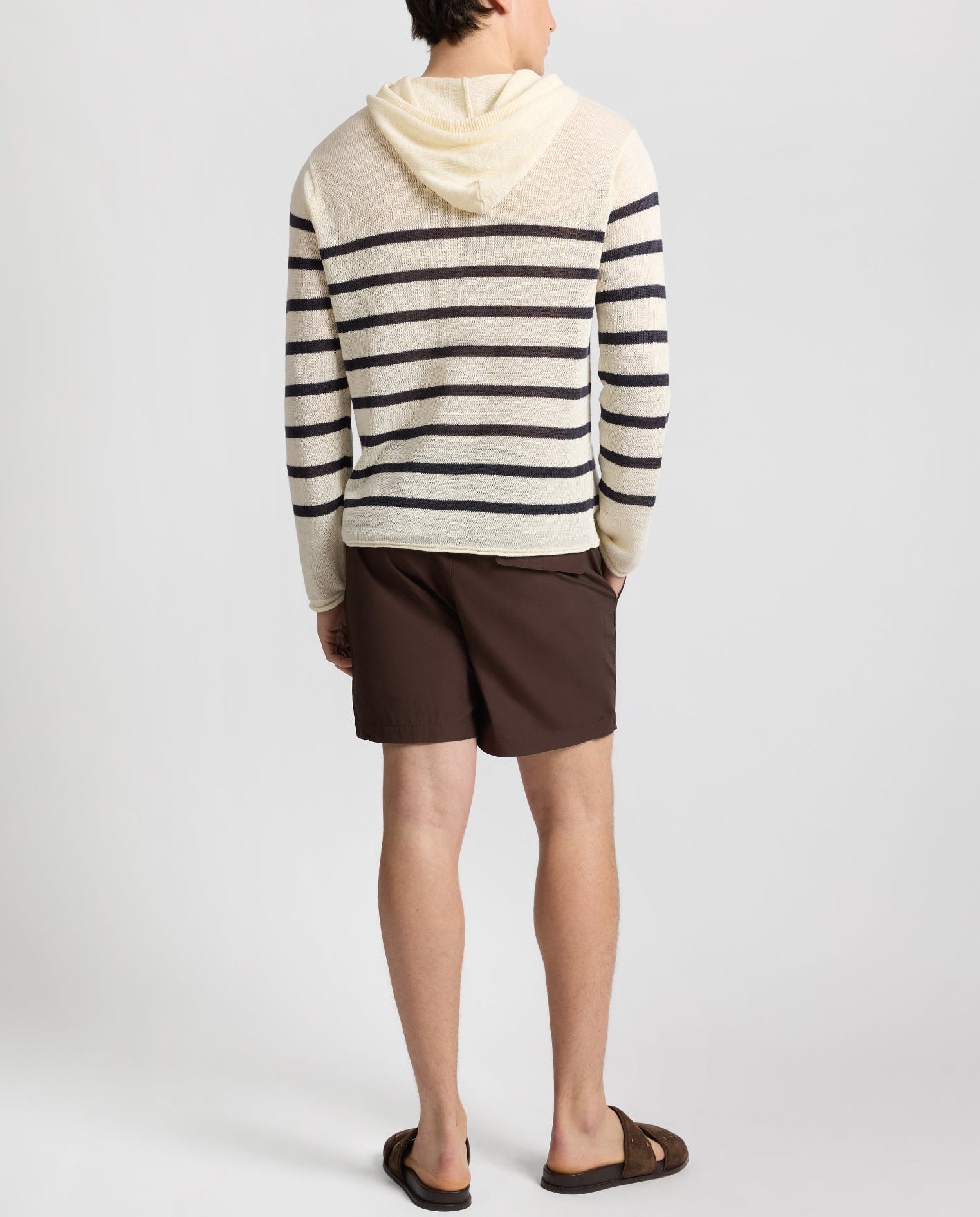 A person stands facing away against a plain background, wearing the Onia Frank Hooded Sweater—a cream and black striped breathable knit—paired with brown shorts and brown sandals.
