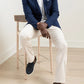 A man sits on a wooden chair wearing the Peter Millar Excursionist Flex Blazer, a white polo, light beige pants, and dark slip-on shoes in a minimal, light-colored room.