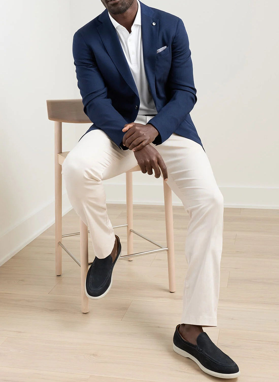 A man sits on a wooden chair wearing the Peter Millar Excursionist Flex Blazer, a white polo, light beige pants, and dark slip-on shoes in a minimal, light-colored room.