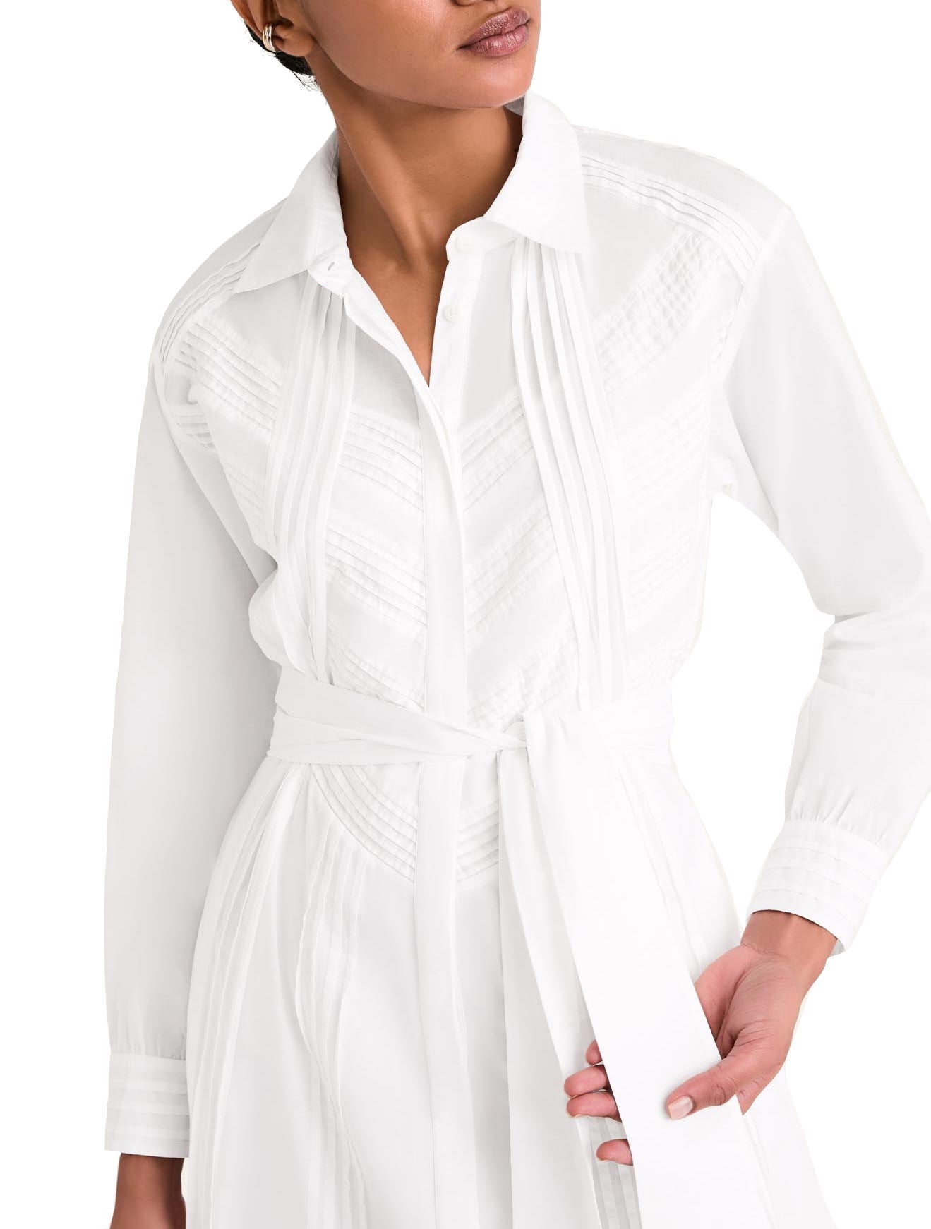 A person wearing the Merlette Liberty Dress, a white long-sleeved shirt dress made of Pima cotton from Merlette New York, complete with a collar and matching waist tie, holds the belt while looking to the side.