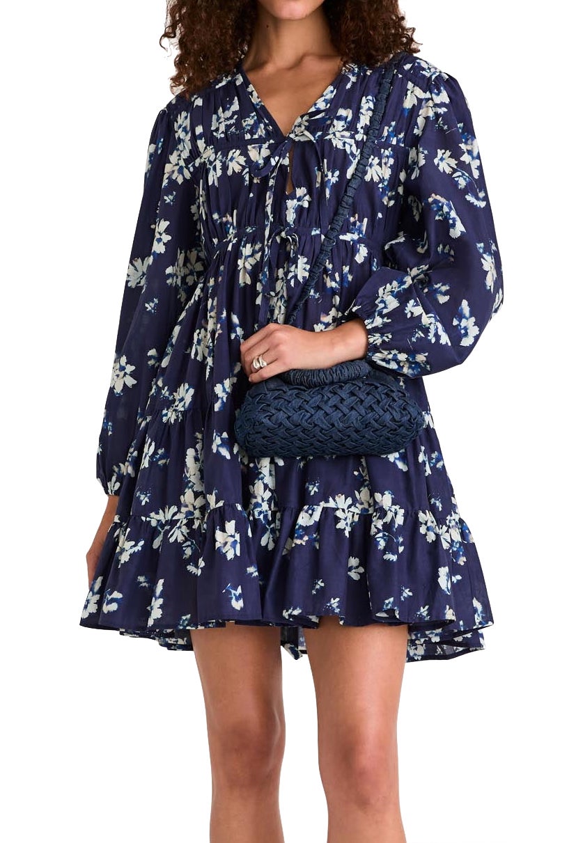 A woman is wearing a navy blue Merlette Lasse Indigo Floral Dress from Merlette New York and holding a dark blue woven clutch.