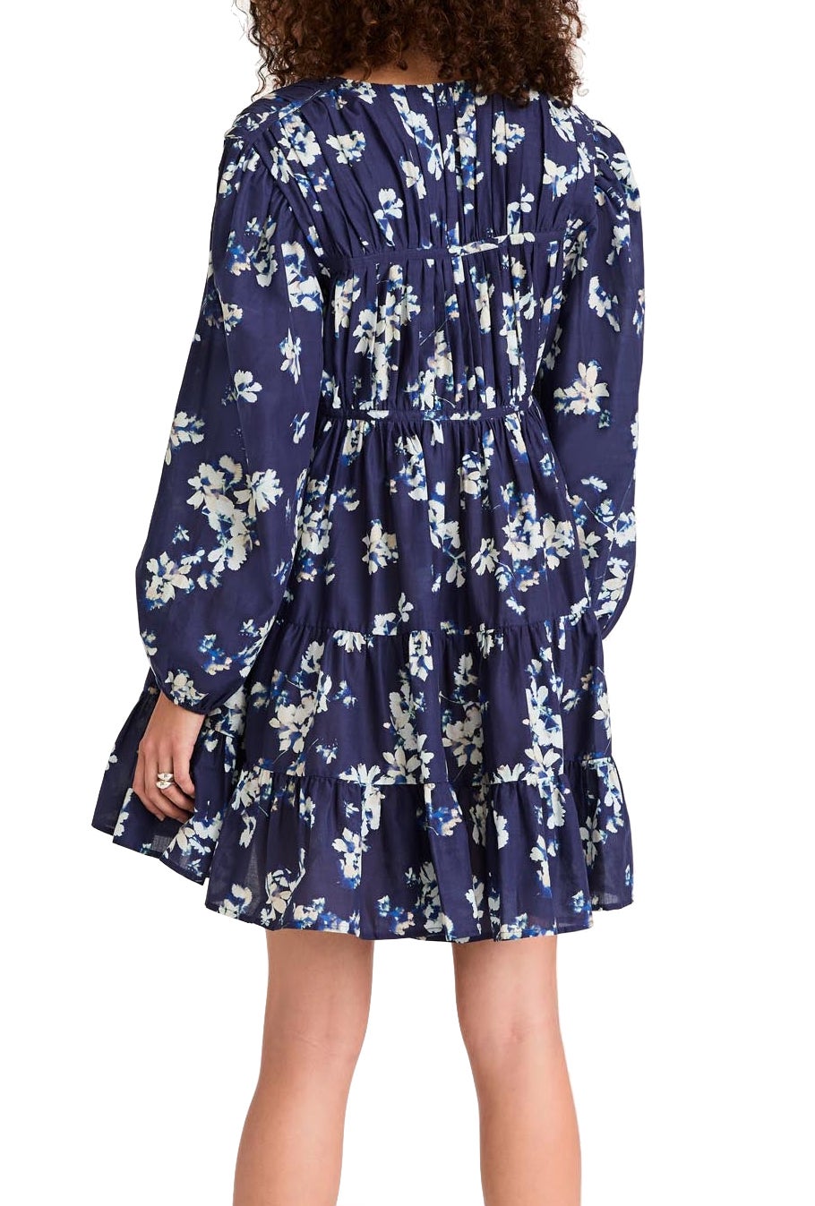 A woman with curly hair is wearing a Merlette Lasse Indigo Floral Dress by Merlette New York. The dress, made from sustainable Pima cotton, has a loose fit and ruffled hem, and she is seen from the back.