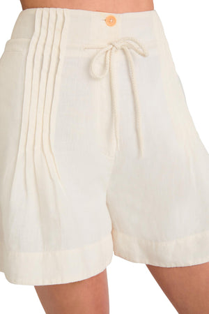 Close-up of a person wearing the Merlette Matin Short by Merlette New York, featuring white linen shorts with pintuck detailing, a drawstring waist, and a single button and zipper closure.