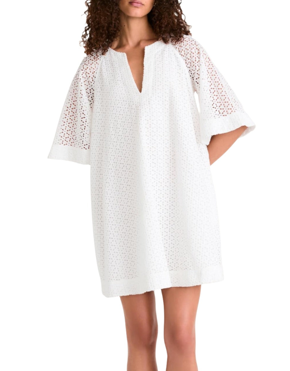 A woman models the Merlette Verdant Dress by Merlette New York—a white cotton eyelet shift dress with three-quarter sleeves and a V-neckline—standing with her hands behind her back.