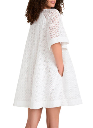 A woman with curly hair wears the loose-fitting, white Merlette Verdant Dress by Merlette New York, featuring cotton eyelet fabric, short sleeves, and pockets, standing with one arm partially visible inside a pocket.