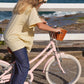 A child wearing a yellow shirt and jeans rides a pink Banwood Kids’ Vintage Classic Bike with a basket near a white fence and the ocean.