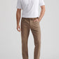 A man stands wearing a white short-sleeve polo, Peter Millar eb66 Performance Five-Pocket Pants in tan, with a comfort waistband, brown belt, and brown sneakers against a light gray background.
