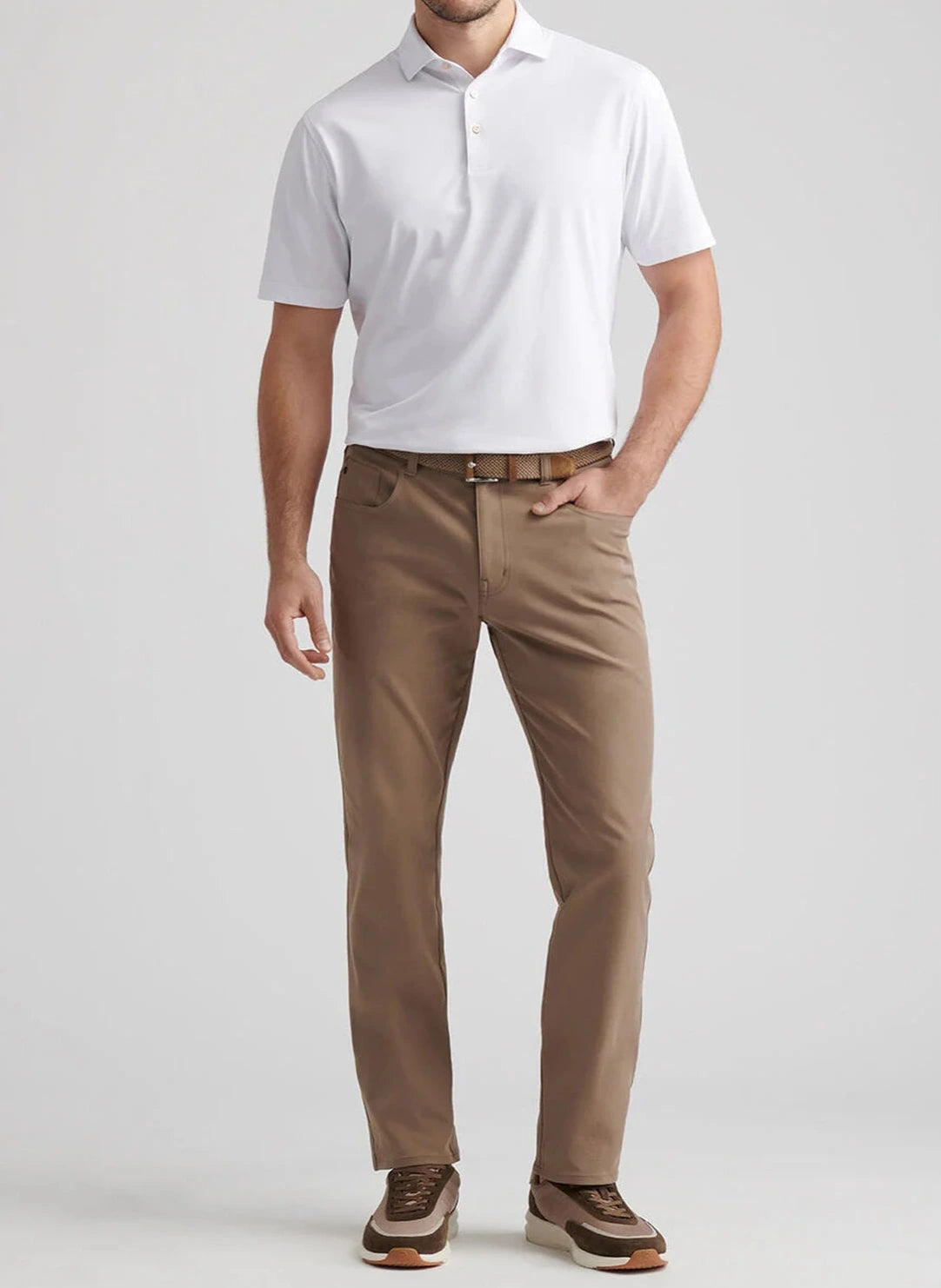 A man stands wearing a white short-sleeve polo, Peter Millar eb66 Performance Five-Pocket Pants in tan, with a comfort waistband, brown belt, and brown sneakers against a light gray background.