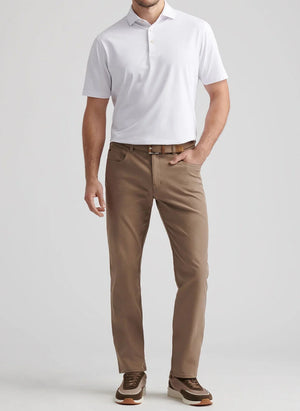 A man stands wearing a white short-sleeve polo, Peter Millar eb66 Performance Five-Pocket Pants in tan, with a comfort waistband, brown belt, and brown sneakers against a light gray background.