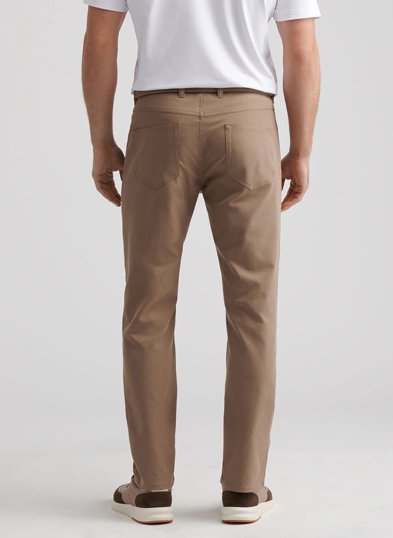 A man faces away, wearing a white polo and Peter Millar eb66 Performance Five-Pocket Pant made from stretch twill with a comfort waistband, paired with brown-and-white shoes against a plain background.