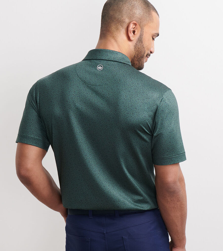 A man faces away, wearing a dark green Peter Millar No Slow Play Performance Jersey Polo with four-way stretch and blue pants, set against a plain light background.