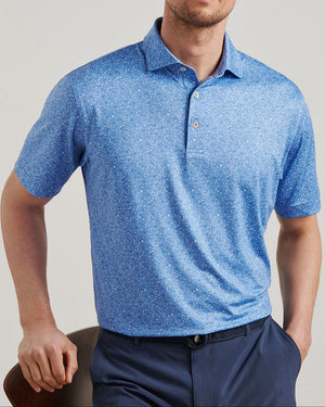 A man wearing the Peter Millar Champagne and Caviar Performance Jersey Polo with four-way stretch and a subtle white pattern, paired with navy blue pants, sits on a chair against a plain background.