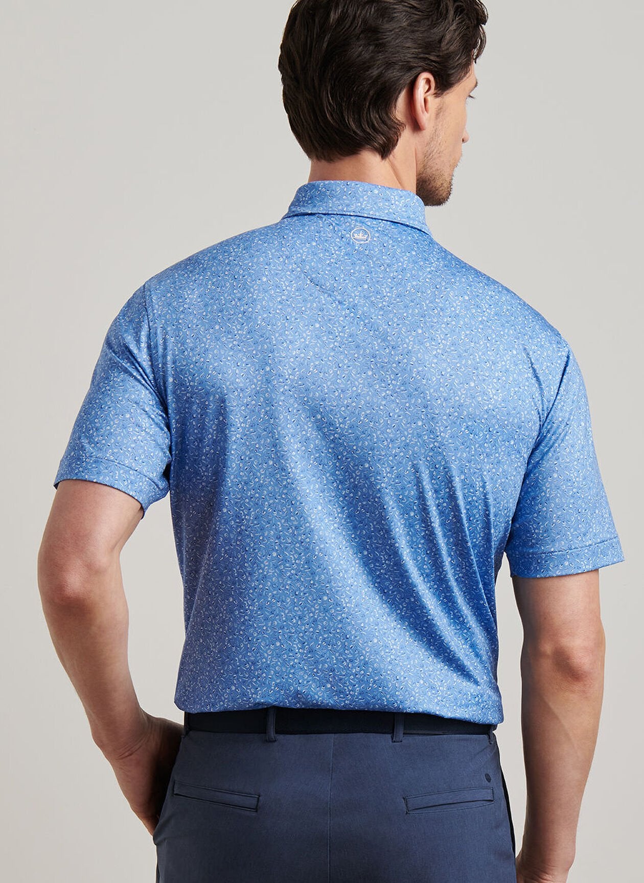 A man with short brown hair wears a light blue Champagne and Caviar Performance Jersey Polo by Peter Millar and dark blue pants, shown from the back against a neutral background.