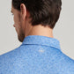 A man seen from behind wears the Peter Millar Champagne and Caviar Performance Jersey Polo—a light blue patterned shirt with a small white logo below the collar, designed for four-way stretch and breathable comfort.