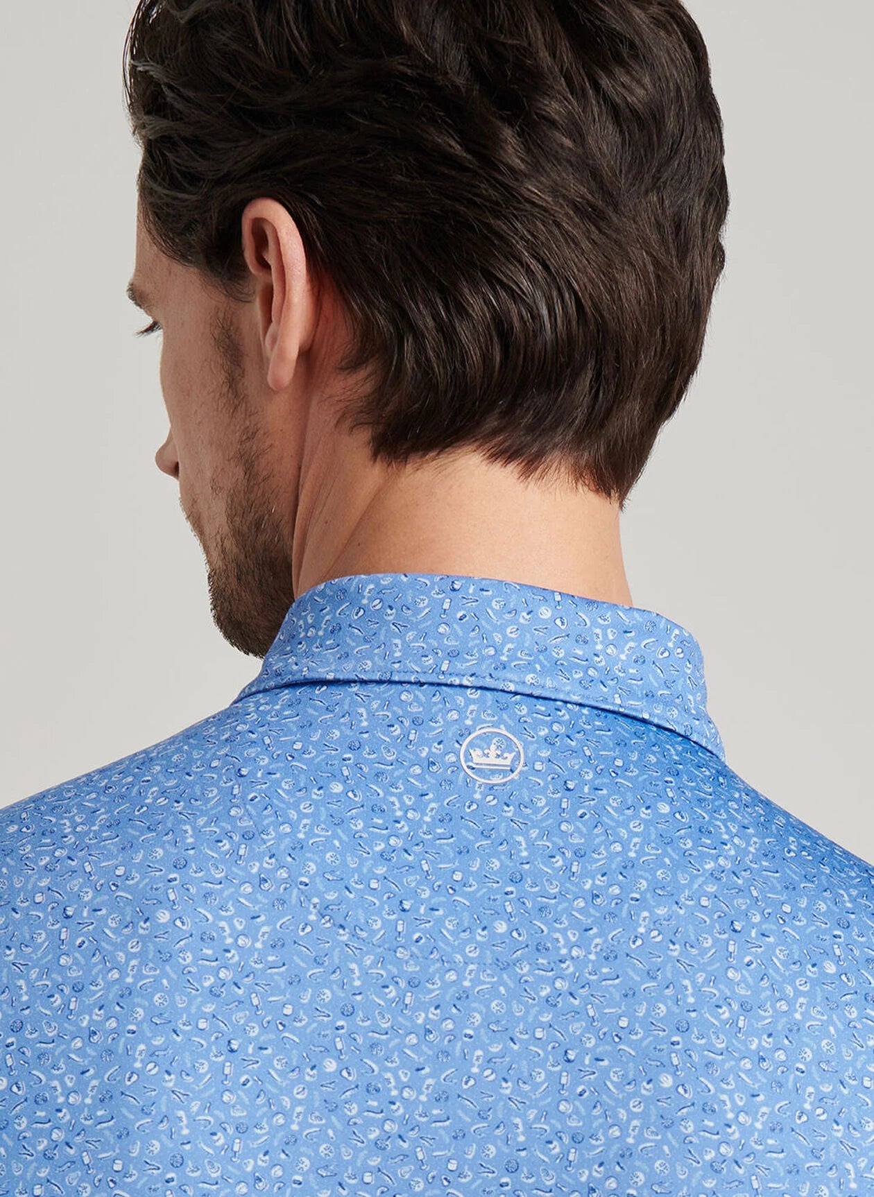 A man seen from behind wears the Peter Millar Champagne and Caviar Performance Jersey Polo—a light blue patterned shirt with a small white logo below the collar, designed for four-way stretch and breathable comfort.