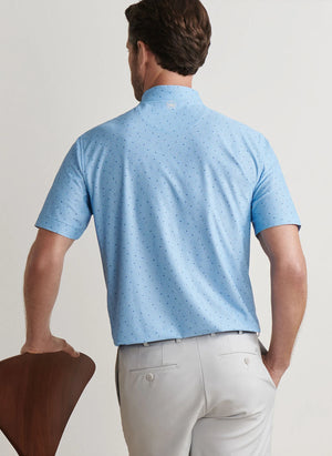 A man in a light blue Peter Millar Old Fashioned Performance Mesh Polo with a subtle dot pattern and light gray pants stands facing away, resting one hand on the back of a wooden chair.