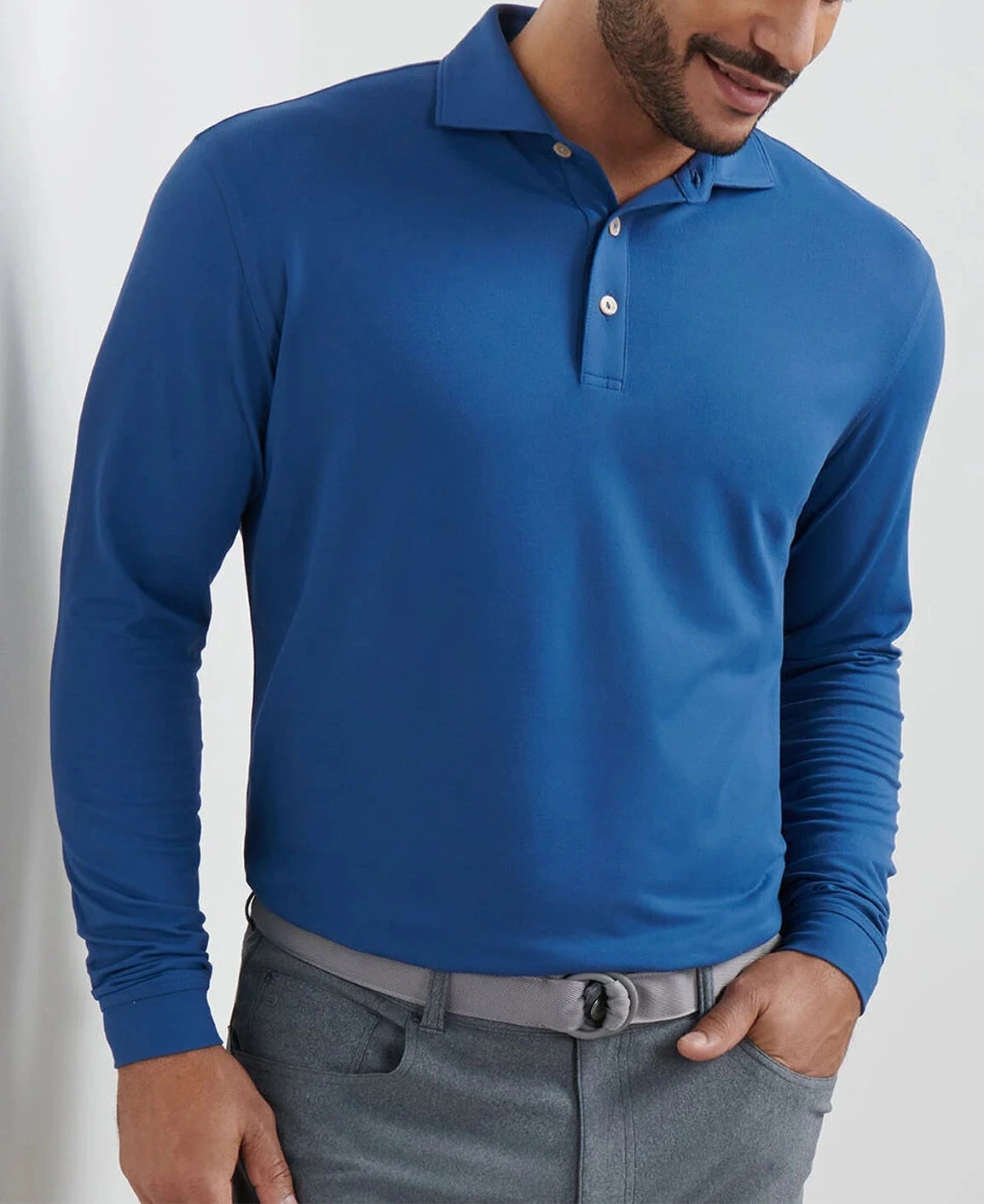 A man wears the Peter Millar Lyons Performance Jersey Long-Sleeve Polo in blue, featuring UPF 50+ sun protection, tucked into gray pants with a gray belt, standing against a light background.