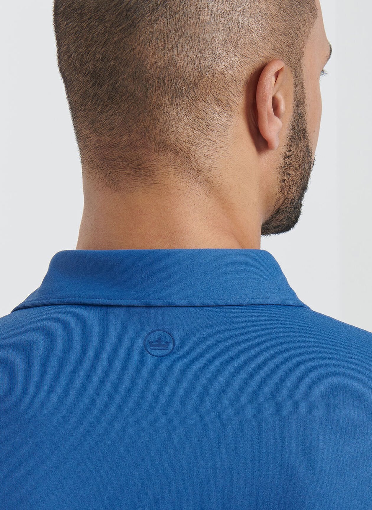 A man shown from behind wears the Peter Millar Lyons Performance Jersey Long-Sleeve Polo in blue, featuring a small circular crown logo below the collar and UPF 50+ sun protection for comfort outdoors.