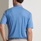 A man faces away from the camera, wearing a light blue Peter Millar Featherweight Mélange Performance Polo and beige pants against a plain background.