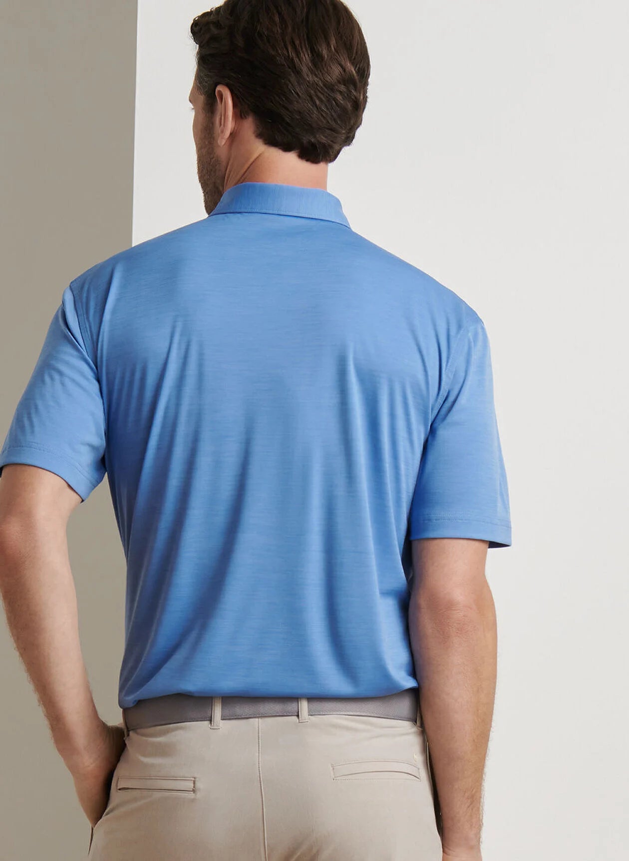 A man faces away from the camera, wearing a light blue Peter Millar Featherweight Mélange Performance Polo and beige pants against a plain background.
