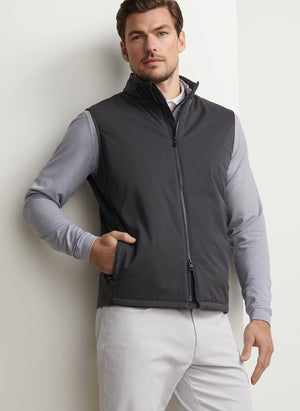 A man stands against a white wall wearing the Peter Millar Fuse Hybrid Vest—a gray zip-up vest with an insulated front panel—over a light gray long-sleeve shirt and light pants, one hand in his vest pocket.