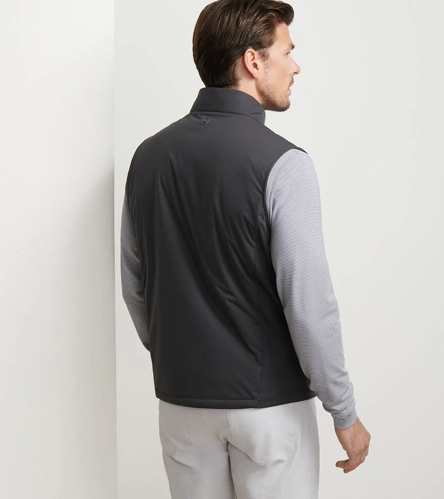 A man stands facing away, wearing a Peter Millar Fuse Hybrid Vest in dark gray over a light gray long-sleeve shirt and white pants, set before a plain light-colored wall.