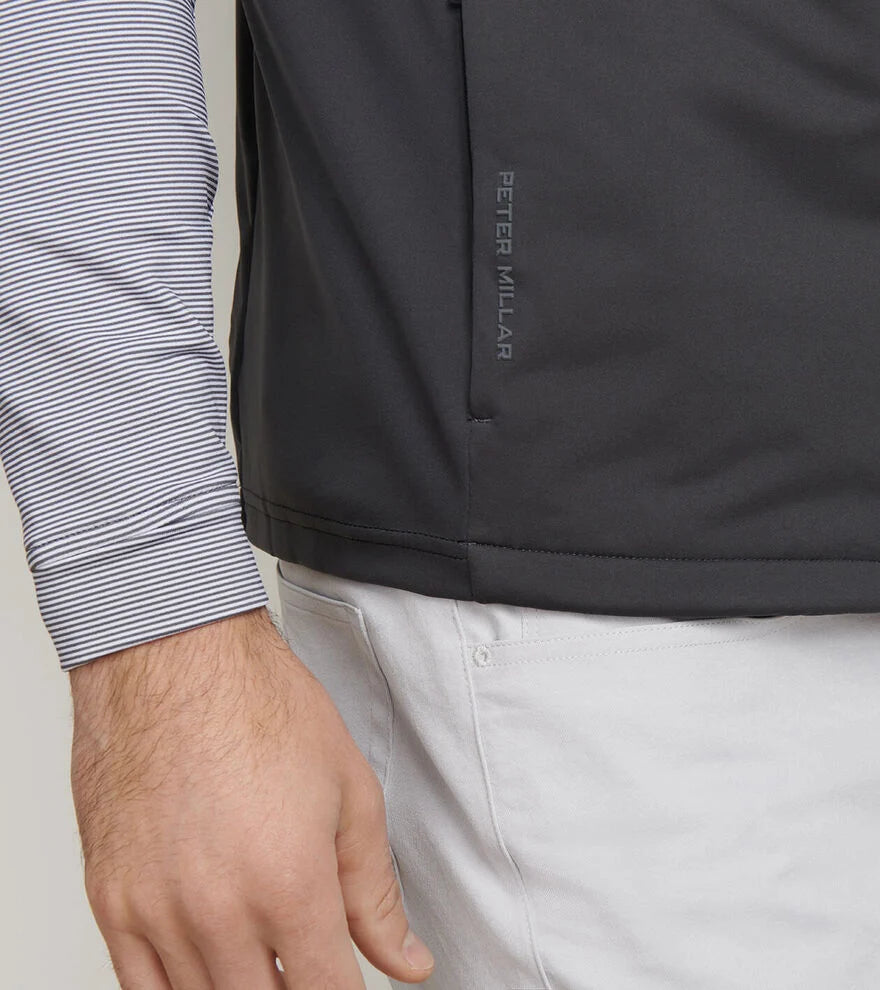 A person wearing a dark Peter Millar Fuse Hybrid Vest, a striped shirt, and white pants.
