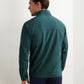 A man stands facing away, wearing the Peter Millar Dunes Half-Zip in teal performance fabric and navy pants, against a plain white wall.