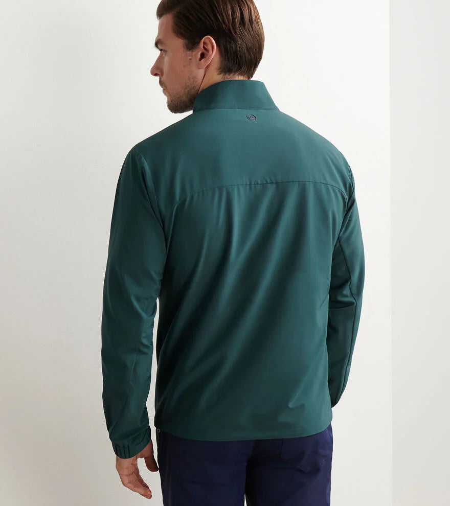 A man stands facing away, wearing the Peter Millar Dunes Half-Zip in teal performance fabric and navy pants, against a plain white wall.