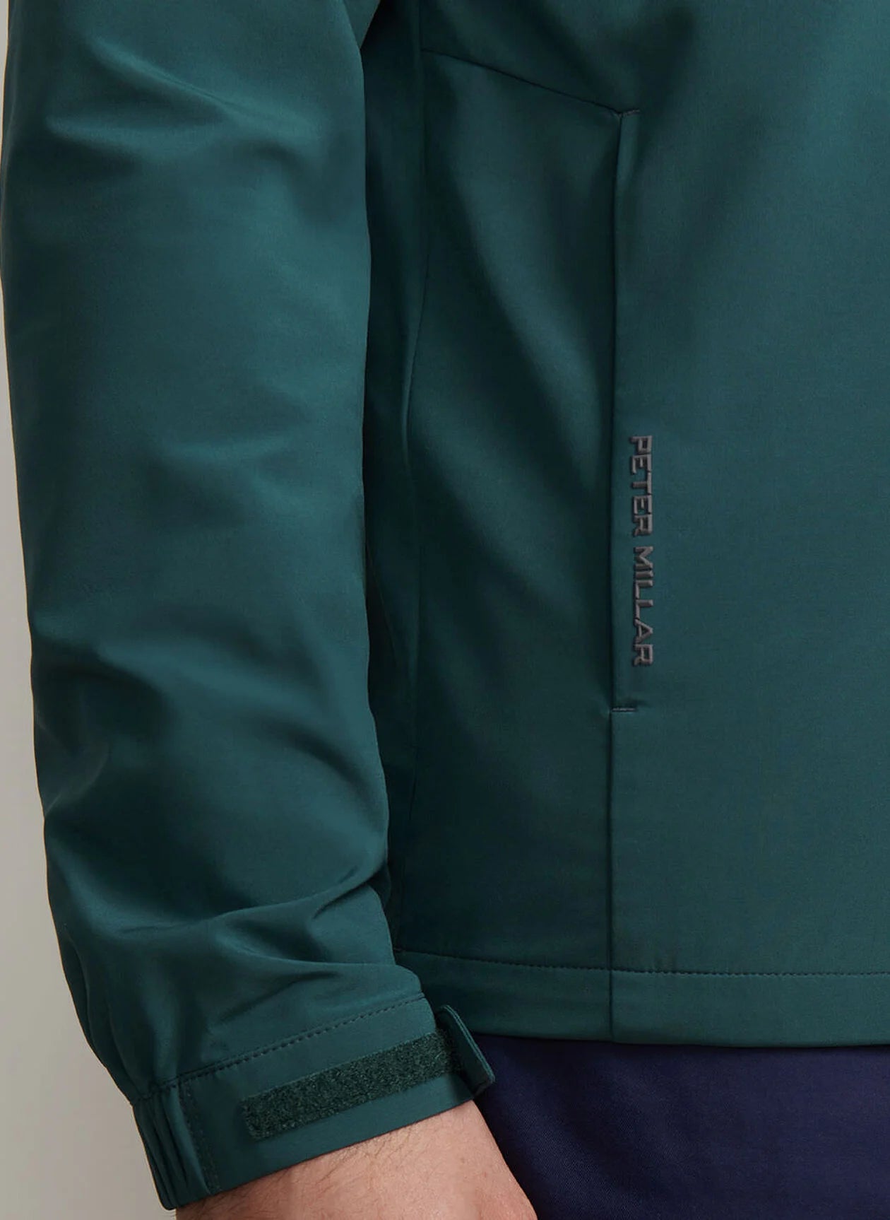 Close-up of a person's arm wearing the Peter Millar Dunes Half-Zip in teal, showcasing four-way stretch double-weave fabric, a visible logo, side pocket, and a velcro strap at the cuff.