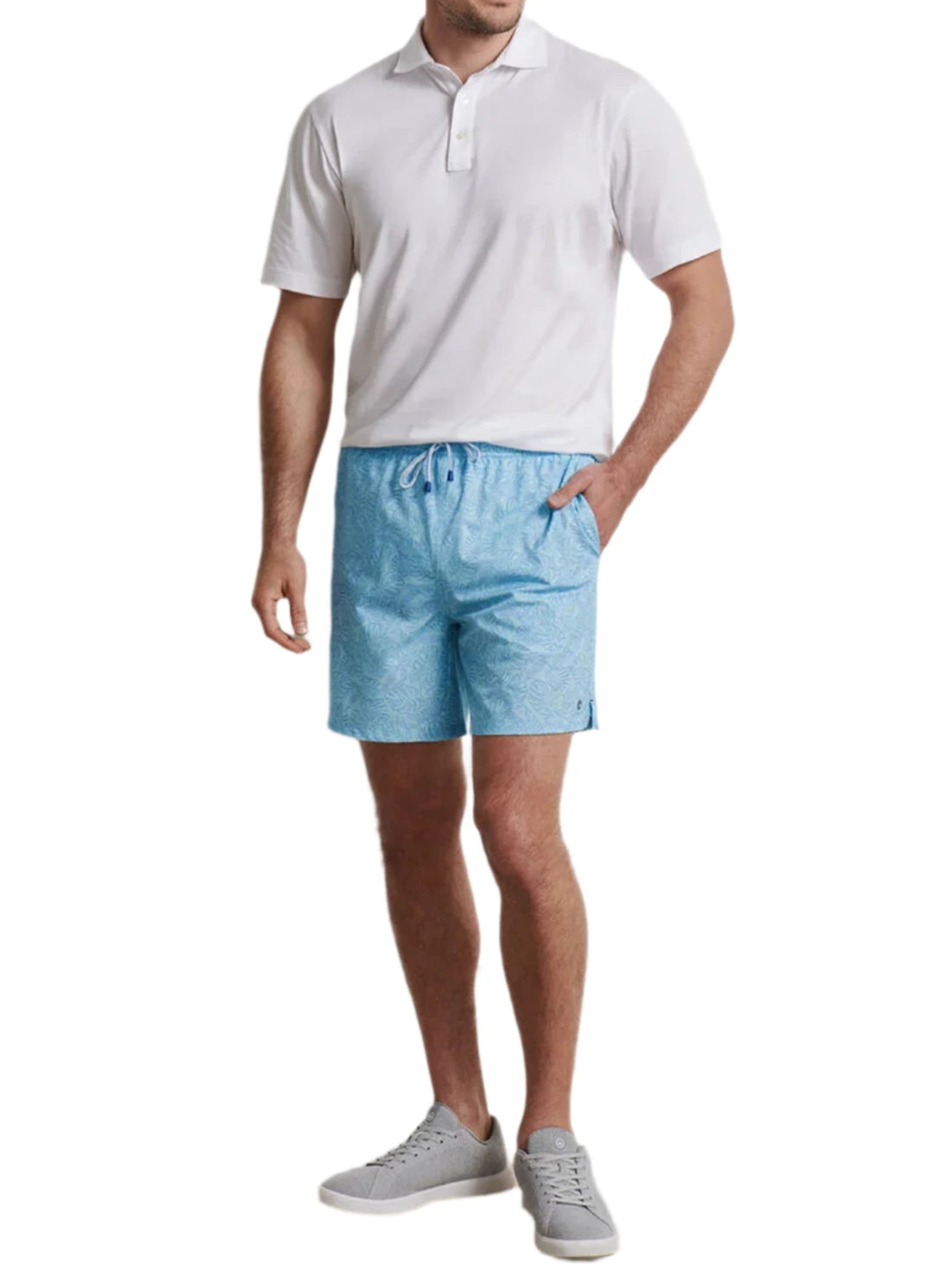 A man stands with one hand in his pocket against a white background, wearing a white polo shirt, light blue Peter Millar Tropics Swim Trunk, and gray sneakers.