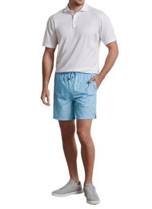 A man stands with one hand in his pocket against a white background, wearing a white polo shirt, light blue Peter Millar Tropics Swim Trunk, and gray sneakers.