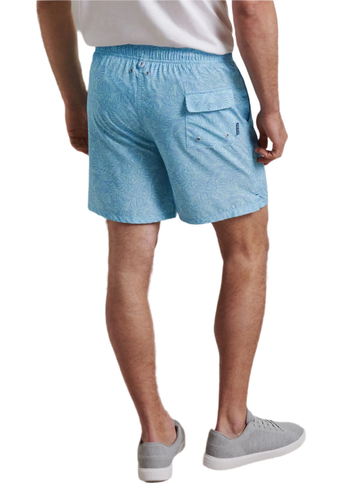 A man is shown from the back wearing a white t-shirt, Peter Millar Tropics Swim Trunk with light blue palm tree print and four-way stretch, and gray slip-on shoes.
