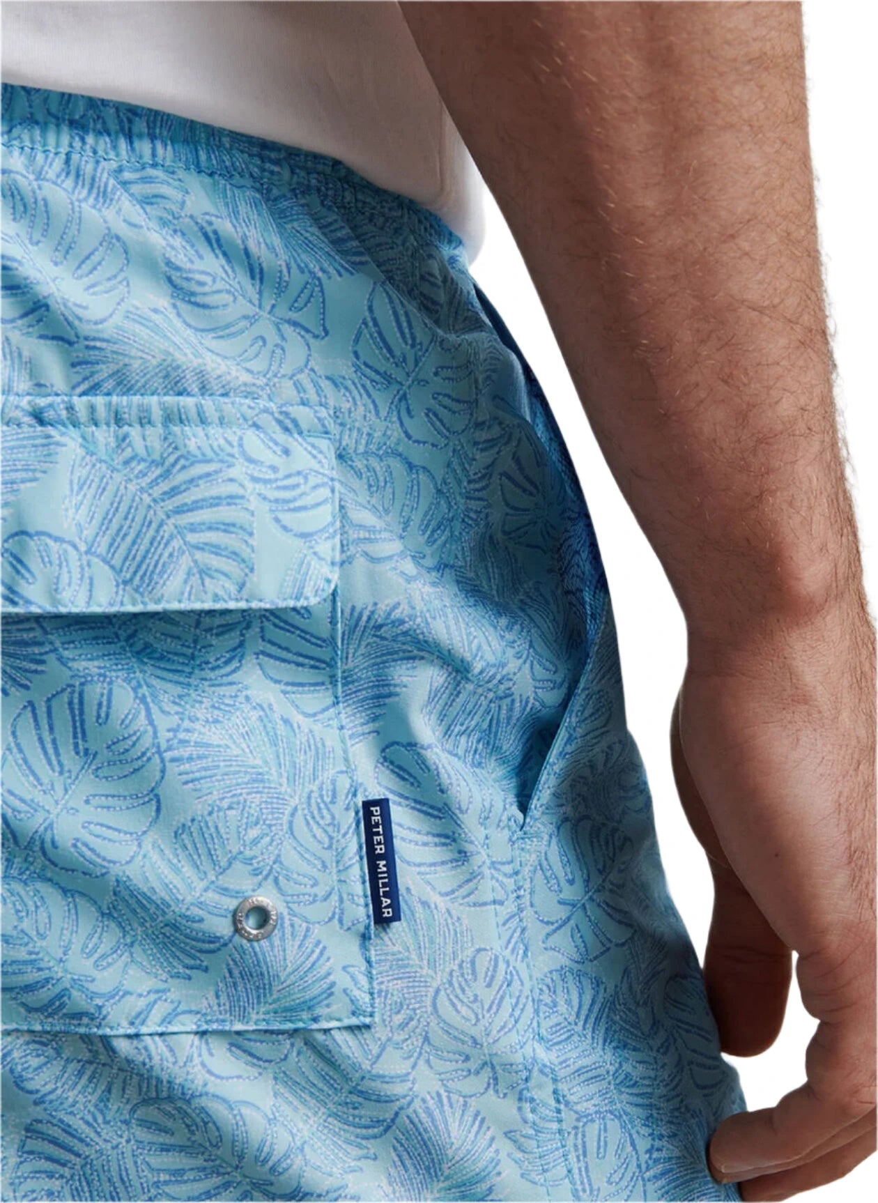 A close-up of a person wearing Peter Millar Tropics Swim Trunk, featuring a light blue palm tree print, back pocket, and brand label.