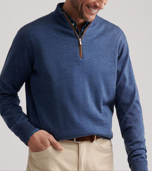 Man wearing the Peter Millar Autumn Crest Suede Trim Quarter-Zip in blue over a collared shirt and beige pants, smiling while looking down with one hand in his pocket.