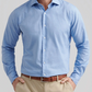 A man wears the Peter Millar Highlands Soft Twill Cotton Sport Shirt in light blue, featuring a spread collar, tucked into beige pants with a brown belt, standing with hands in pockets against a plain background.