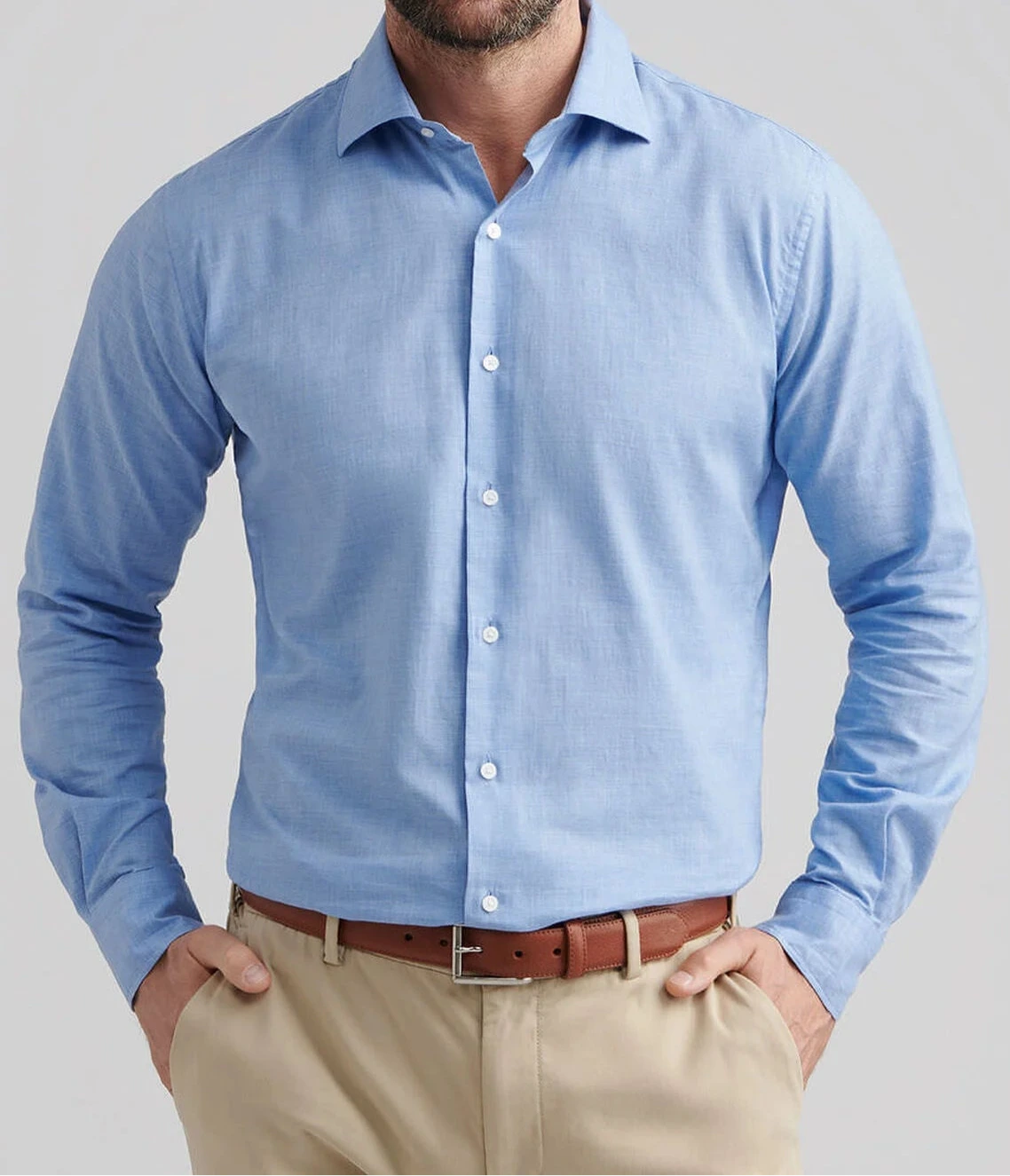 A man wears the Peter Millar Highlands Soft Twill Cotton Sport Shirt in light blue, featuring a spread collar, tucked into beige pants with a brown belt, standing with hands in pockets against a plain background.