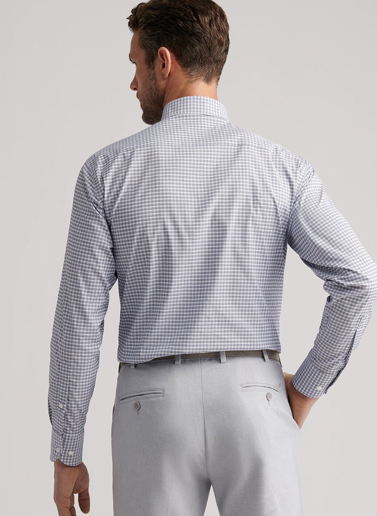 A man stands with his back to the camera, wearing a Peter Millar Cashiers Performance Twill Sport Shirt in light blue check, tucked into light grey dress pants. This moisture-wicking shirt blends comfort and style for any occasion.