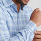 A man wearing the Peter Millar Bar Harbor Performance Flannel Sport Shirt in light blue and white plaid adjusts his sleeve, with only the lower half of his face showing.