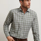 A man wears the Peter Millar Whitecap Performance Flannel Sport Shirt, a long-sleeve green and gray plaid shirt made from lightweight performance fibers, as he looks to his right against a plain background.