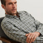 A man with short hair and stubble wears a Peter Millar Whitecap Performance Flannel Sport Shirt while sitting in a gray cushioned chair, looking slightly to the side with a neutral expression.