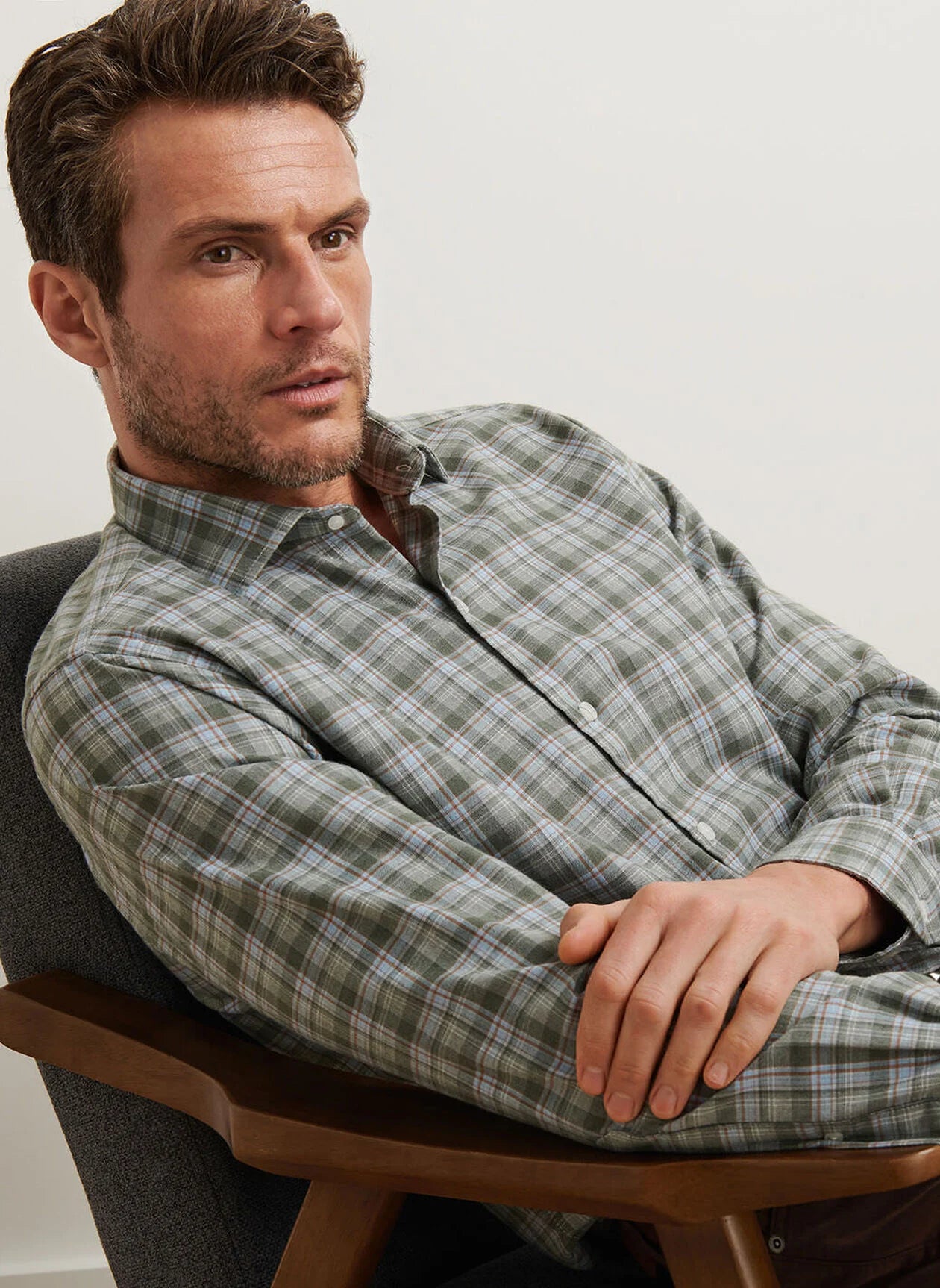 A man with short hair and stubble wears a Peter Millar Whitecap Performance Flannel Sport Shirt while sitting in a gray cushioned chair, looking slightly to the side with a neutral expression.