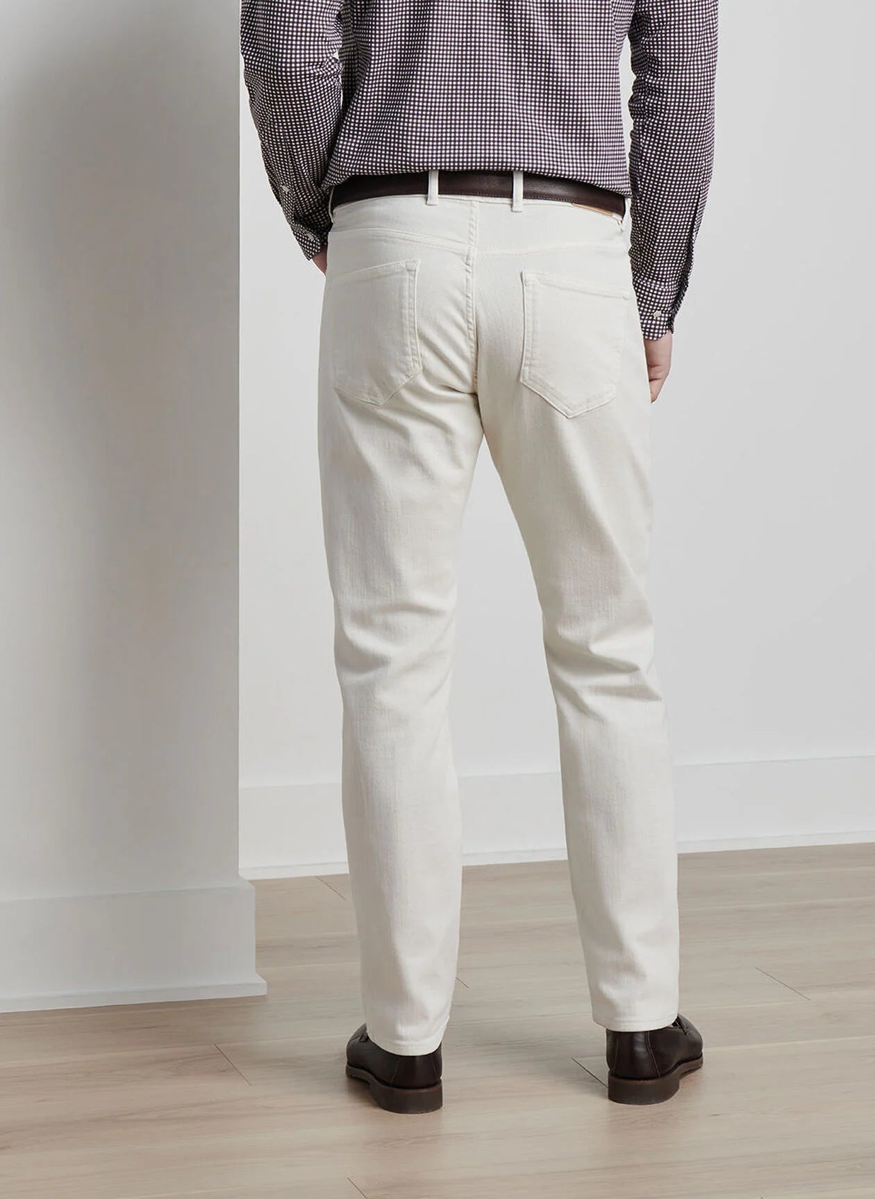 A person wearing a checkered shirt, white Peter Millar Fairfield Garment Dyed Denim pants, and brown shoes stands with their back to the camera, facing a wall.
