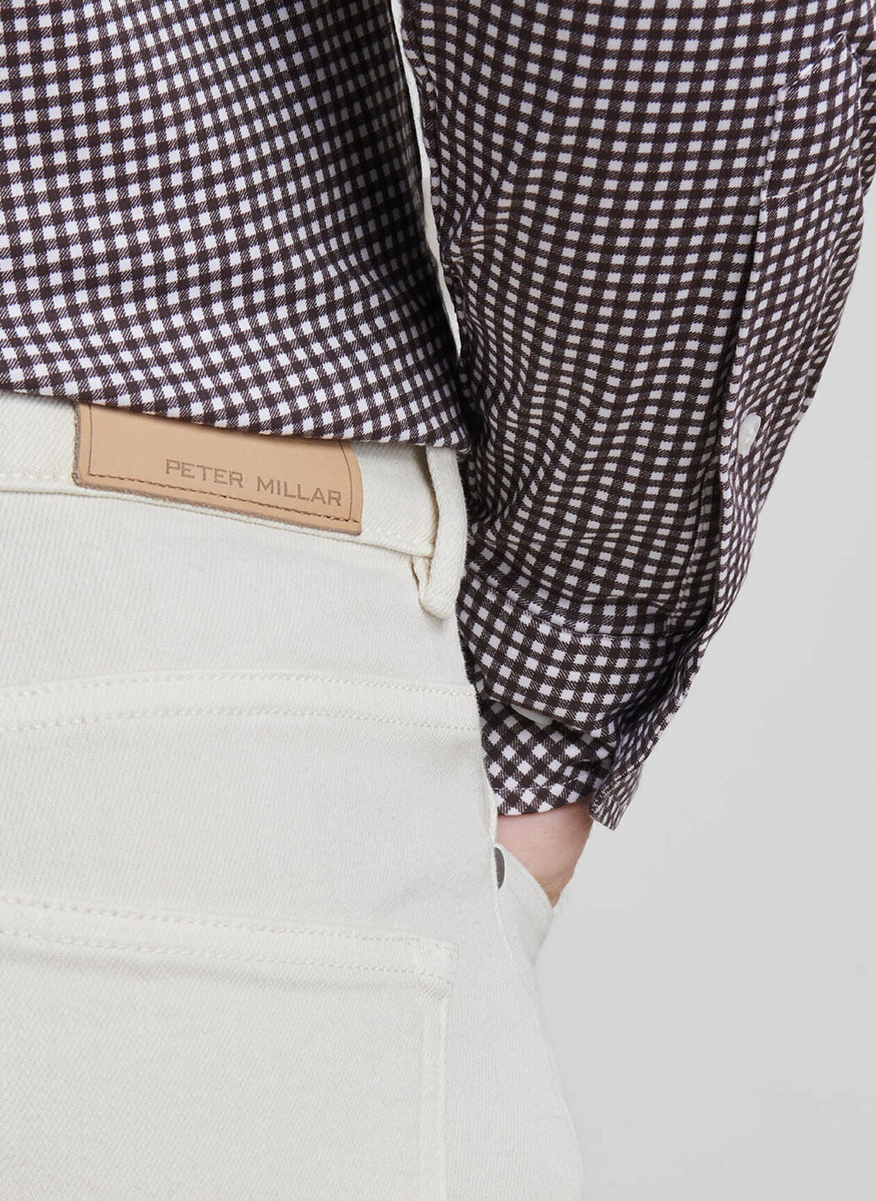 Close-up of a person wearing the Fairfield Garment Dyed Denim by Peter Millar, featuring a brown and white checkered shirt tucked into the cream-colored jeans with a visible Peter Millar leather label above the back pocket.