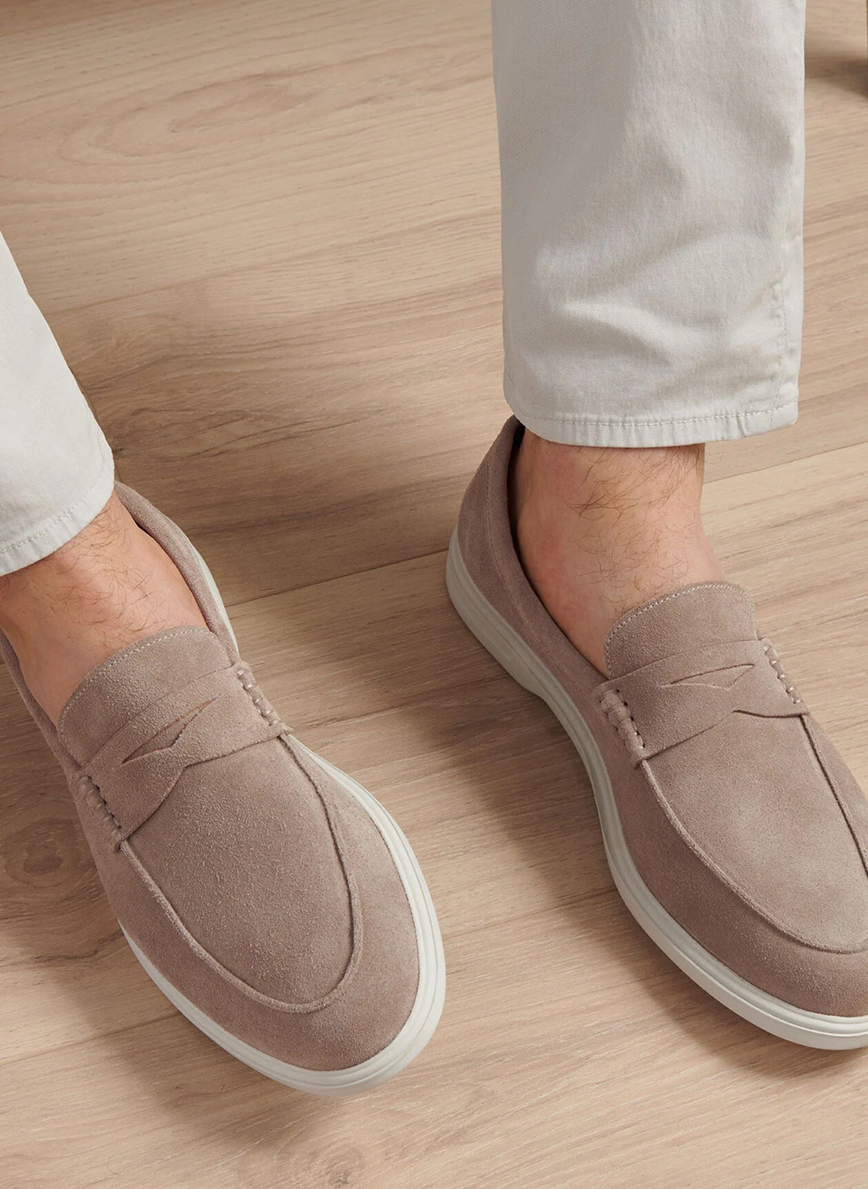 A person wearing Peter Millar Excursionist Penny Loafers, featuring water-resistant suede and lightweight EVA outsoles, pairs them with light-colored pants while standing on a light wooden floor.