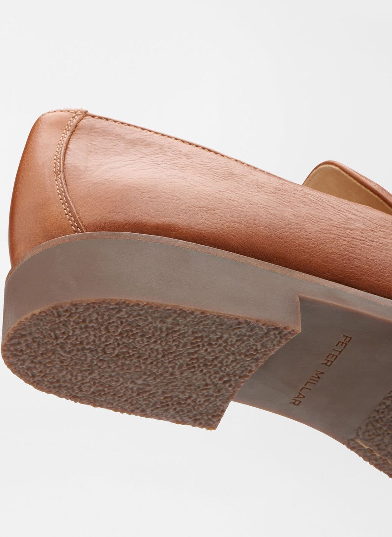Close-up of the Madison Penny Loafer by Peter Millar, crafted from brown tumbled calf leather with a textured rubber sole, displayed side and bottom view against a white background—a modern and versatile shoe.