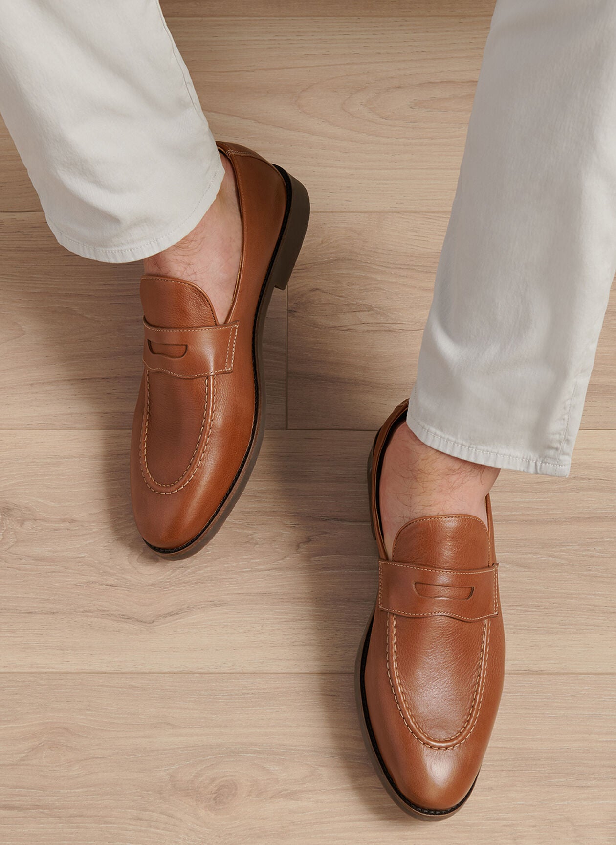 A person in light-colored pants wears Peter Millar's Madison Penny Loafers, crafted from tumbled calf leather, standing on a light wood floor and displaying modern versatility.