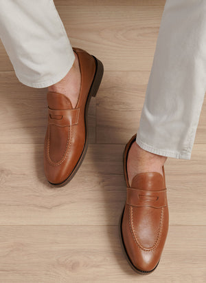 A person in light-colored pants wears Peter Millar's Madison Penny Loafers, crafted from tumbled calf leather, standing on a light wood floor and displaying modern versatility.